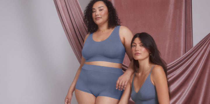 Bianca stands on the left wearing the Cloud Cotton Scoop Bralette and Highwaist Boyshort in Slate, posing confidently against a soft pink draped backdrop. On the right, Anna sits wearing the Cloud Cotton Triangle Bralette in Slate, with a relaxed expression as she rests her arm against.