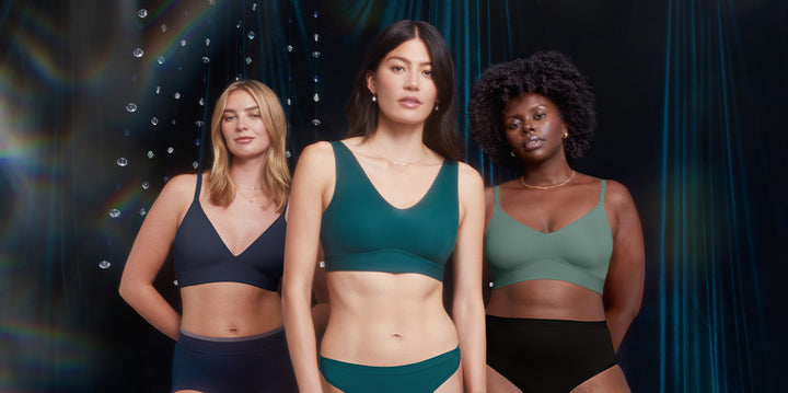 Three models stand confidently in front of a dark, velvet backdrop adorned with hanging crystals. On the left, Megan wears the Bliss Triangle Bralette in Navy. In the center, Anna models the Bliss Bralette in Everglade. On the right, Akesha showcases the Bliss Triangle Bralette in Rainforest paired with black briefs.