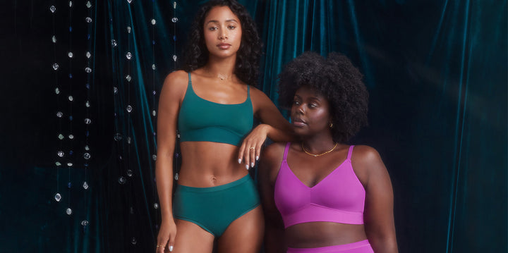 Ally, wearing the Bliss Scoop Bralette and Highwaist Brief in Everglade, rests her arm on Akesha’s shoulder. Akesha, dressed in the Bliss Triangle Bralette in Berry, looks off-camera as they pose against a dark velvet backdrop with hanging crystals.